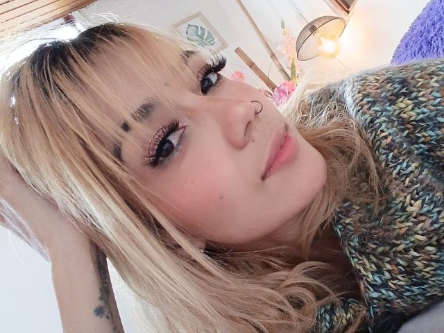 Veroo69, com cabelo loiro, olhos azuis e um piercing no nariz, usando um cachecol verde e marrom tricotado, olhando para a câmera com um leve sorriso..