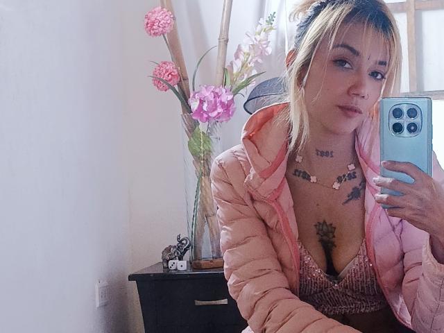 Veroo69 con capelli biondi, giacca gonfia rosa e reggiseno rosa, che si scatta un selfie allo specchio, mostrando tatuaggi e fiori rosa sullo sfondo. Il seno è visibile nel reggiseno..