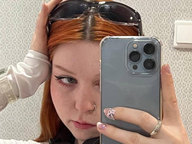 LilliamChevalier with red hair, holding a phone in front of her face, wearing sunglasses on her head, nose ring, and white long-sleeve shirt, with pink manicured nails.