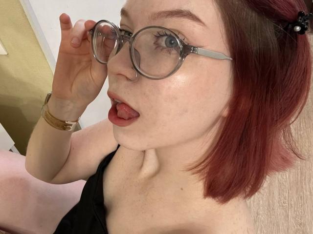 LilliamChevalier with red-dyed hair, wearing glasses, sticking out tongue, and holding glasses up. She has fair skin, a black top, and a gold watch.