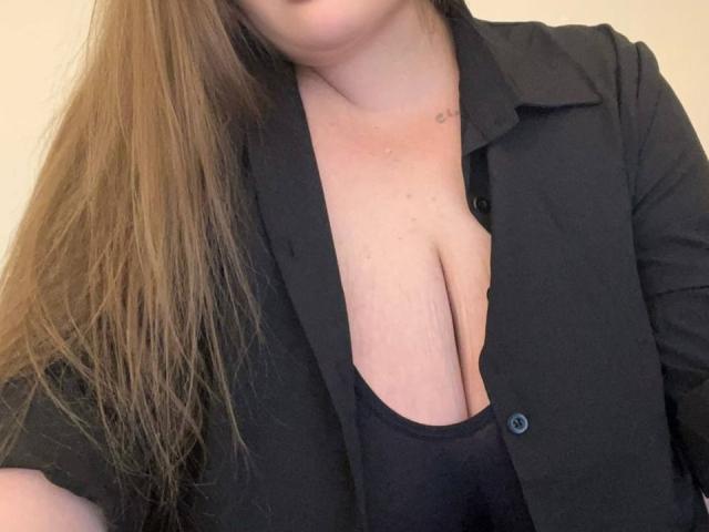 DevilishMuse with long brown hair, black shirt open to reveal large, natural, saggy tits, wearing a black top underneath. Skin is fair with a small mole on her chest.