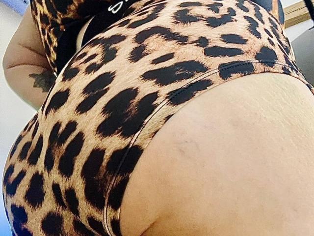 DevilishMuse's ass in a tight, leopard-print dress, showing a tattoo on the left side and a slight bruise on the right. The focus is on her curvy, tan buttocks.
