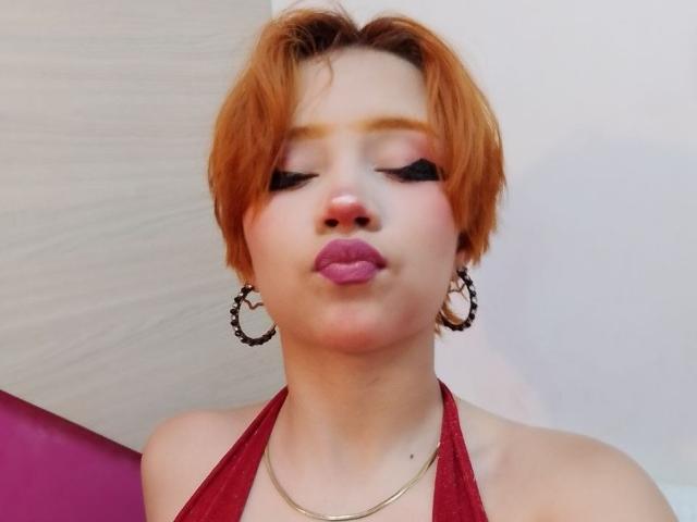 Chloee Williams, com cabelo curto e laranja, delineador preto marcante, batom rosa, lábios rosados e volumosos, vestindo um top vermelho de alças, um colar dourado e grandes brincos de argola, fazendo beicinho..