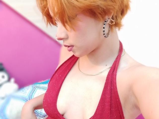 Chloee Williams, con i capelli corti e arancioni, indossa un top rosso scintillante che mette in mostra il seno, ha orecchini a forma di stella e una collana d'argento, e guarda in basso con un'espressione accigliata..