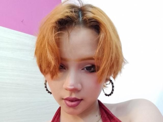 Chloee Williams, met een oranje bob, draagt oorringen, een rode haltertop en roze lippenstift, en kijkt naar beneden met licht open lippen. Haar achterkant is niet zichtbaar..
