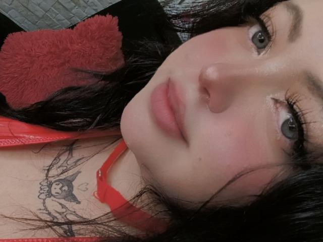 OnixDamon's face with pale skin, green eyes, and black hair, wearing a red top, has a tattoo under her chin with a cute face design, lying down.