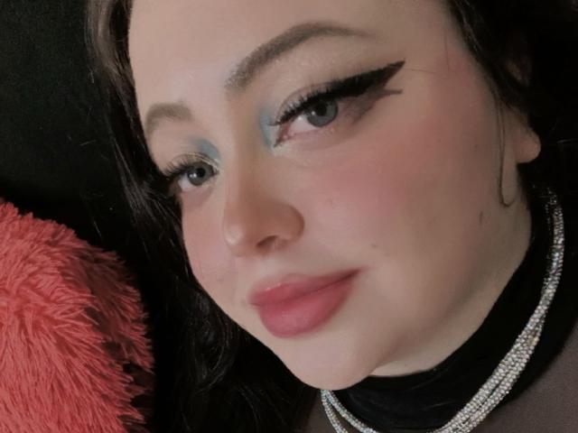 OnixDamon's face with blue eyeshadow, black winged eyeliner, pink lips, fair skin, and a small black nose. She's wearing a black top and a sparkly necklace.