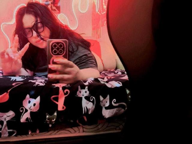 OnixDamon with glasses, dark wavy hair, grey shirt, lying on black cat print bed, making a peace sign, taking selfie with pink phone, red light in background.