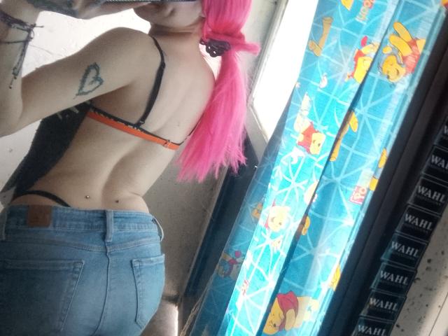 Madiy69 with pink hair, black bra, and blue jeans, showing her ass and a small tattoo on her back. Mirror selfie, reflection in a window with colorful curtains.