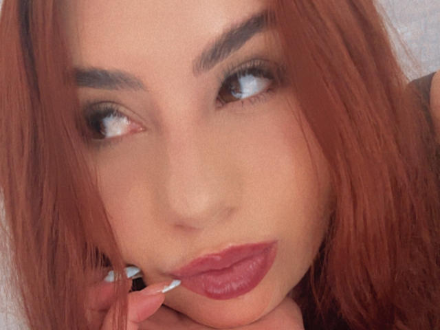 VenusxMusa com cabelo ruivo, sobrancelhas escuras, olhos azuis e batom rosa, olhando para o lado, tocando os lábios com um dedo com unhas brancas..