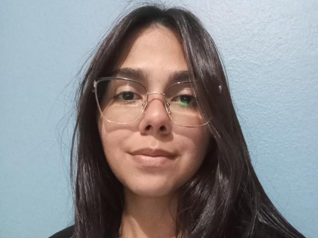 MorenitaBellas com cabelos longos e escuros, usando óculos de grau, tem olhos verdes e castanhos, sorrindo sutilmente, parede azul clara ao fundo. Seios não visíveis, foco no rosto..