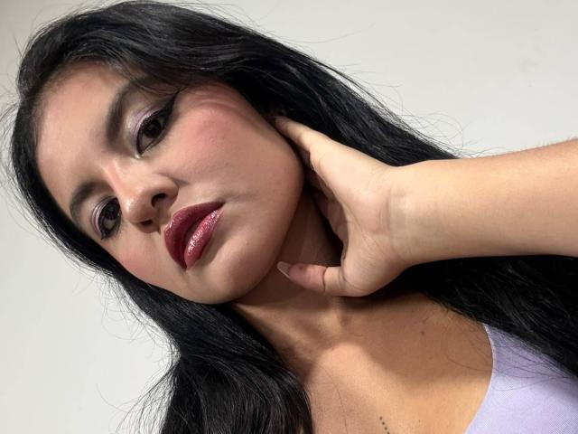 AlessiaVeega with long black hair, red lipstick, and pink eyeshadow, touching her cheek with one hand, wearing a lilac top, showing a slightly pouty expression.