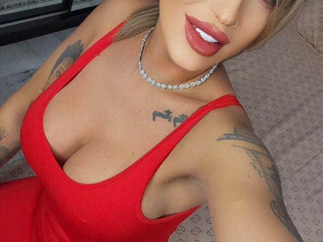 NicolSNaya com um vestido vermelho, mostrando seios grandes, braços tatuados, um piercing no nariz e batom vermelho, usando um colar de diamantes, em um selfie..