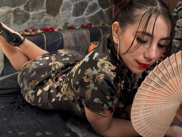 FreyaraVenwo laying on her stomach, wearing a dark floral qipao, holding a fan, with red lipstick, black heels, and her ass raised, looking down with a slight smile.