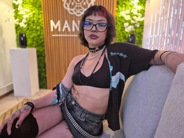 JadeMilleer with short brown hair, glasses, black bra, open shirt, denim shorts, black stockings, tattoos, and jewelry, sitting on a couch, looking at camera.