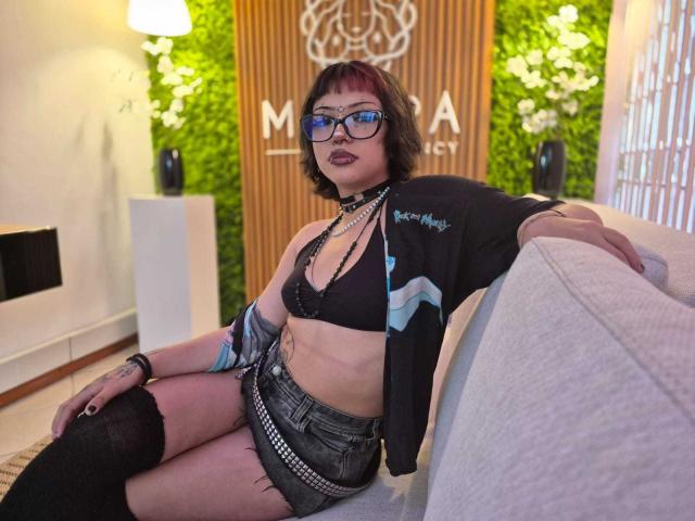 JadeMilleer with short brown hair, glasses, black bra, denim shorts, black jacket, thigh-high socks, sitting on couch, purple lipstick, tattoos, necklace, and arm warmers, looking at camera.