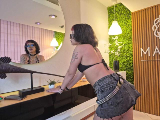 JadeMilleer with shoulder-length brown hair, black bra, and denim skirt, leaning on a counter, showing her back tattoo, looking in a mirror, with glasses and dark lipstick.