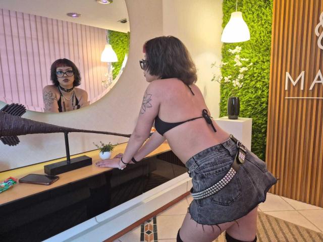 JadeMilleer with tattoos, black bikini top, and denim skirt, leaning on counter, looking in mirror, showing her back and ass, wearing glasses, with green wall and pendant lights.