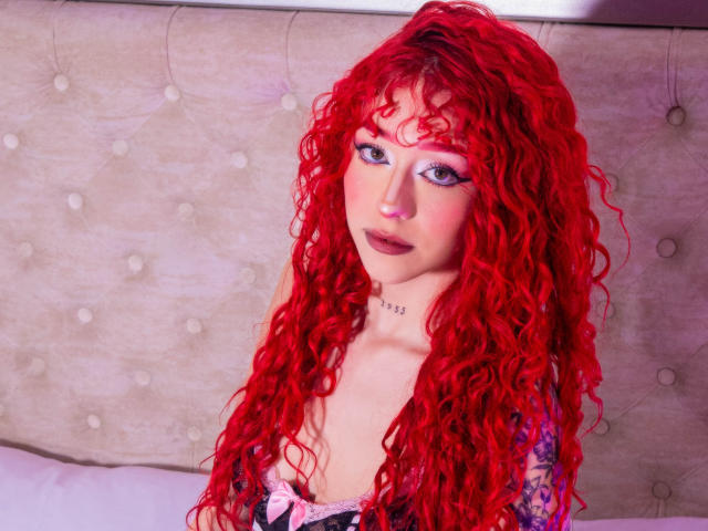 GaiaDoll met felrood krullend haar, roze oogschaduw, paars lippenstift, tatoeages op de rechterarm, gekleed in zwart en roze lingerie, kijkt naar de camera met een lichte glimlach..