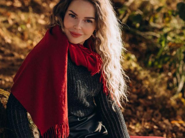 AnnaLoves, avec ses cheveux blonds ondulés, son foulard rouge, son pull noir en tricot et son pantalon noir, est assise à l'extérieur, souriante, avec ses yeux marron clair et un maquillage discret. Elle a l'air mignonne et séduisante..