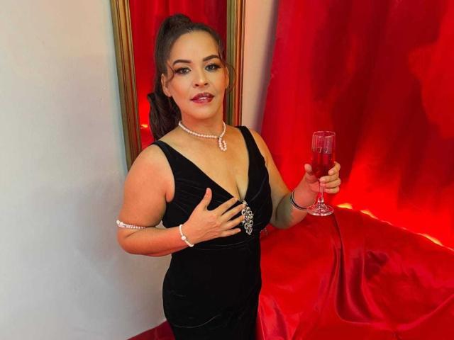 LizethBells with big tits in a black dress, holding a red drink, wearing a pearl necklace and bracelet, standing in front of red curtains, smiling.
