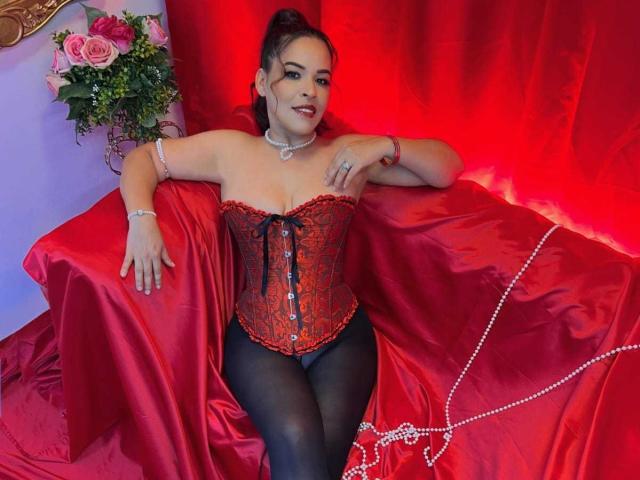 LizethBells with red lace corset, black pantyhose, and red beads, sitting on red satin-covered couch, arms resting, looking at camera, dark hair in ponytail, red lipstick.