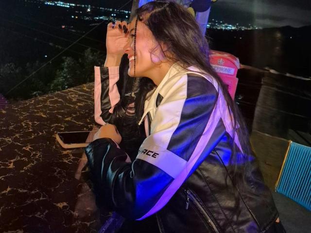 Isabella Voss, com seus longos cabelos escuros, está rindo e cobrindo a boca, vestindo uma jaqueta preta e branca com a inscrição "ICE" na manga, sentada em uma mesa de mármore..