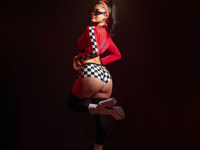MadeleinSimone with big ass, wearing checkered black and white shorts, red top, black mask, white high heels, black thigh-highs, and red headband, standing in dark background.
