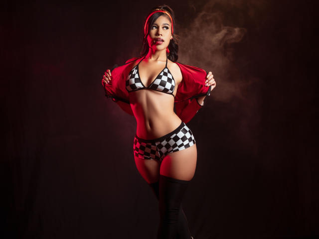 MadeleinSimone con top y pantalones de bikini a cuadros, chaqueta roja y medias negras hasta el muslo, de pie, fondo con humo, sosteniendo la chaqueta abierta, mostrando sus pechos y glúteos, lápiz labial rojo, con confianza..
