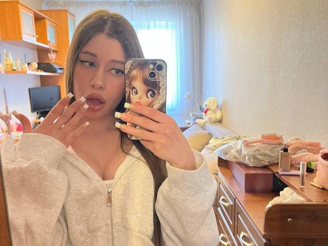 BetseySchwiebert with long brown hair, pouty lips, and white hoodie, holding phone with anime sticker, mirror selfie, showing nails, in cozy room with wooden dresser and stuffed toy.