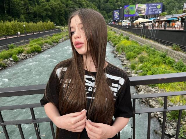 BetseySchwiebert with long brown hair, wearing a black shirt, standing by a river, holding her hair, looking to the side, with a relaxed expression, outside on a bridge.