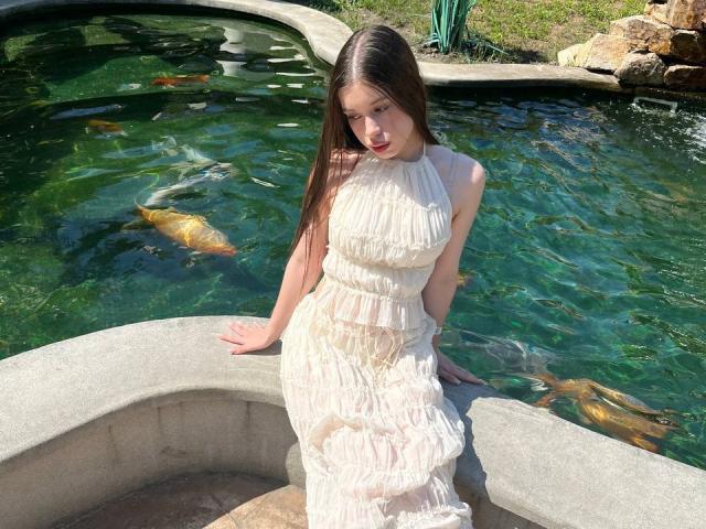 BetseySchwiebert with long brown hair, wearing a sheer white dress, sitting by a clear green pond with visible goldfish, looking down with a slight smile.