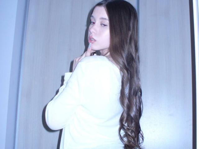 BetseySchwiebert with long, wavy brown hair, white shirt, and finger touching lips, looking back over shoulder, showing her ass. Blue light casts a cool glow.