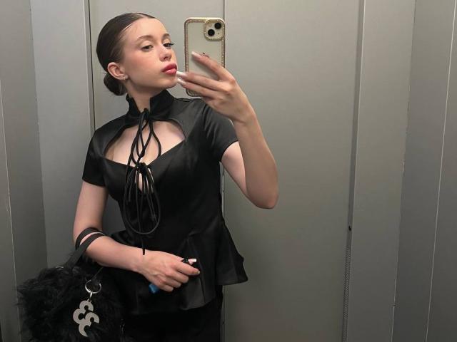 BetseySchwiebert with pouty lips, taking a mirror selfie in a grey elevator. She's wearing a black, cut-out, lace-up top, holding a black bag and a blue item.