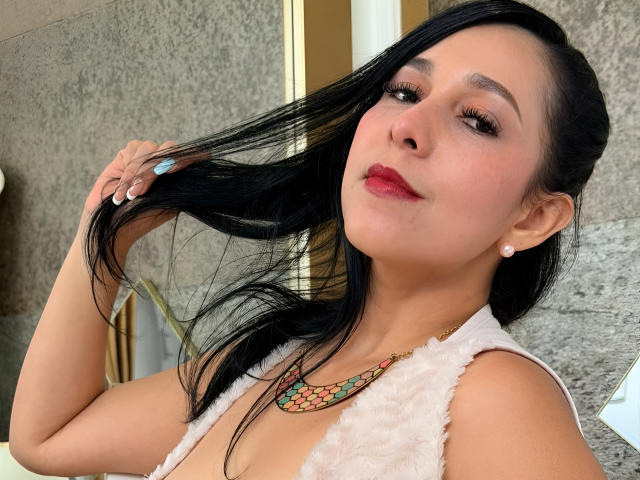 Katty Rivera, avec ses cheveux noirs qu'elle tient attachés, porte un haut blanc en fourrure, des boucles d'oreilles de perles, un collier coloré, du rouge à lèvres rouge, et elle sourit, exhibant sa poitrine, et a des ongles bleus..