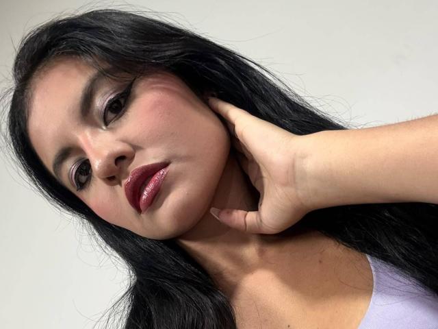 AlessiaVeega has long black hair, pinkish skin, and shiny red lipstick. She's touching her neck with her right hand, wearing a purple top. Her expression is slightly pouty.