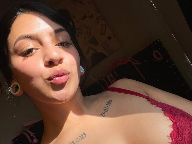 LonxEva with tan skin, black hair, yellow ear gauges, red lace bra, tattoos on chest, pouty lips, sunlight on face, smiling slightly, visible shadow on right.