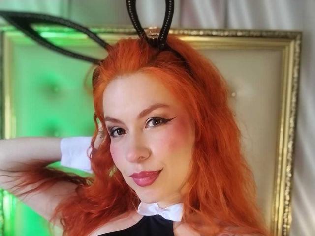 LilithSnop, avec ses cheveux orange, ses cornes de diable noires, son col blanc et son rouge à lèvres rose, souriant à la caméra, les bras derrière la tête, éclairage vert et blanc, mettant en valeur sa poitrine..
