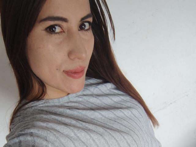 SofiaAnal, com olhos castanhos, pele clara e cabelos castanhos longos, vestindo uma camiseta cinza texturizada, sorrindo para a câmera. Ela tem uma pequena marca de beleza abaixo do olho direito..