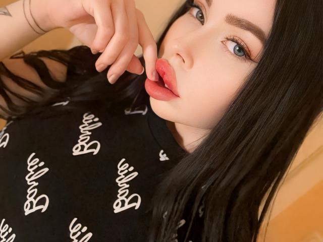 VelvetDoll, avec ses cheveux noirs, ses yeux bleus et ses lèvres roses pulpeuses, touche ses lèvres avec son doigt, porte une chemise noire avec une inscription blanche et a un tatouage au poignet..