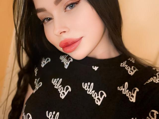 VelvetDoll avec une peau pâle, des yeux bleus et du rouge à lèvres, portant un haut noir avec le mot "will" en blanc, souriant à la caméra, avec de longs cheveux noirs et des lèvres pulpeuses, légèrement retroussées..