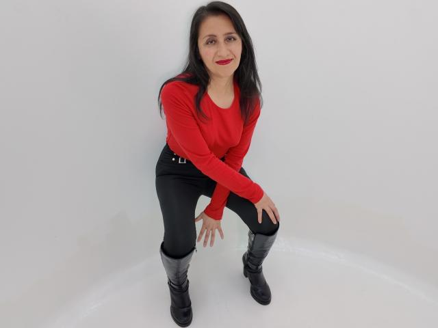 Cyntiia, agachada com as mãos nos joelhos, vestindo uma blusa vermelha de manga comprida, calças pretas e botas pretas, tem batom vermelho e cabelo preto, sorrindo para a câmera..