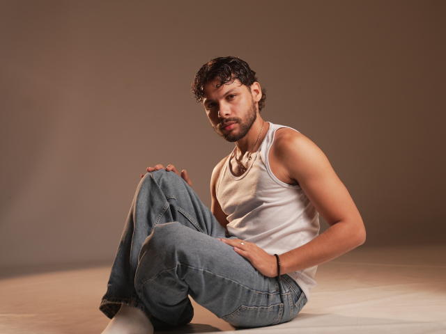 Dmalessandro, a muscular dude with curly brown hair, wearing a white tank top and blue jeans, sits with knees up, hand on knee, looking at camera.