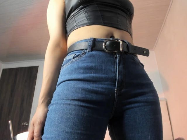 KathyM wearing a black leather crop top and blue jeans with a black belt, showing a slim waist and tight jeans. Her ass is prominent. Low angle shot.