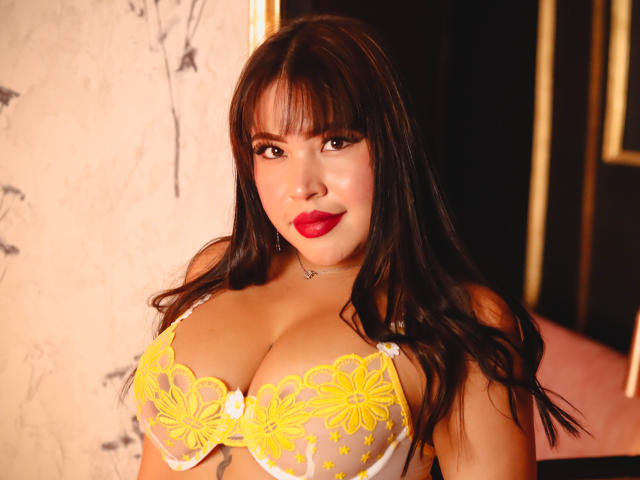 OliviGrace with big boobs in a yellow lace bra, red lipstick, long brown hair, and a cute face, looking directly at the camera with a sexy smile.
