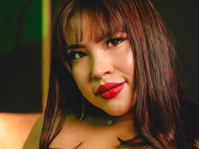 OliviGrace tem cabelo castanho escuro, batom vermelho e um leve sorriso. Ela está usando brincos verdes e tem uma pele brilhante e lisa. Seus seios não estão visíveis na imagem..