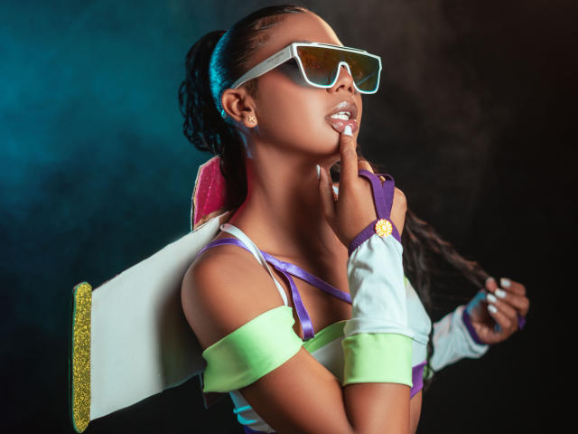 MalloryKim with dark skin, wearing futuristic glasses, green and purple off-shoulder top, holding a white board against her back, finger near mouth, with long braided hair.