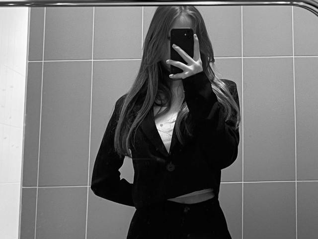 Black-and-white mirror selfie of LanieDistel with long hair, covering face with phone, wearing black blazer, white shirt, and black pants, hand in pocket, showing a bit of midriff.