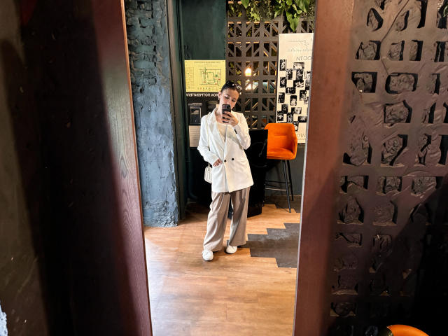 Mirror selfie of LunaNoirX in a white robe, beige pants, and white sneakers, standing with a phone in a dark, textured room with orange chair and wooden floor.