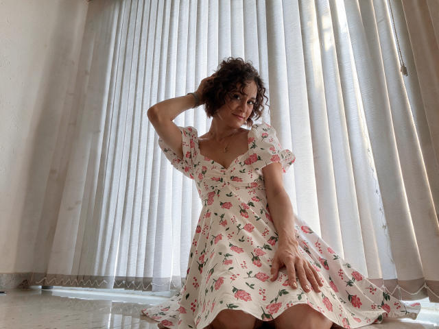 LunaNoirX, con cabello rizado, vestida con un vestido floral, está sentada en el suelo, frente a persianas verticales, con una mano en el cabello, sonriendo, y el vestido ligeramente levantado, mostrando su trasero..